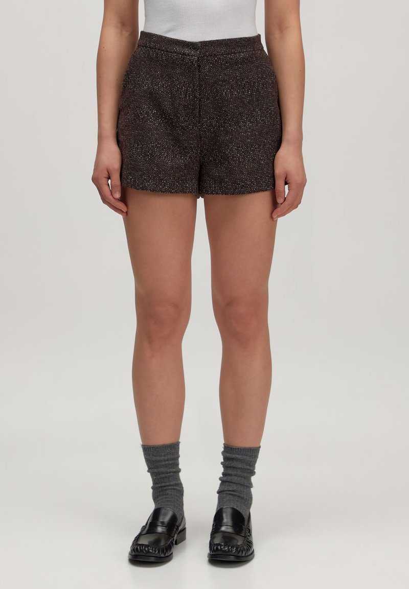 Vrouw in donkerbruin gestructureerde shorts, grijze opgerolde sokken en zwarte leren loafers die tegen een effen achtergrond staat.