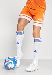 Pantalones cortos de fútbol naranjas con franjas blancas y el logo de Adidas, combinados con calcetas blancas con franjas azules, y botas blancas. Un balón de fútbol multicolor descansa debajo.