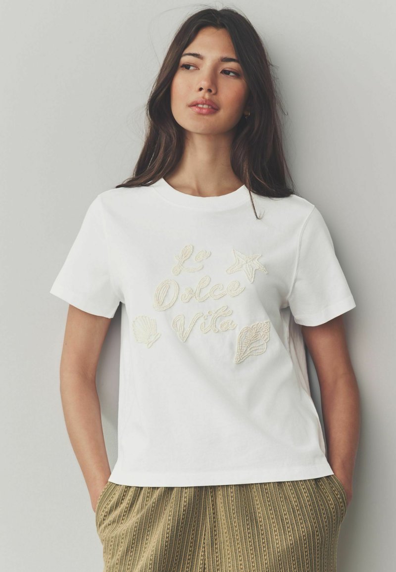 T-shirt en coton blanc avec broderie du texte "La Dolce Vita" accompagné de motifs d'étoile, de coquillage et de feuille. Col rond, manches courtes, coupe décontractée.