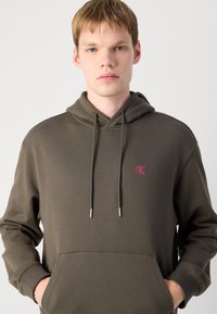 Felpa verde oliva scura con tasca frontale, cappuccio con coulisse e logo rosa sul petto. Il tessuto ha una texture liscia.