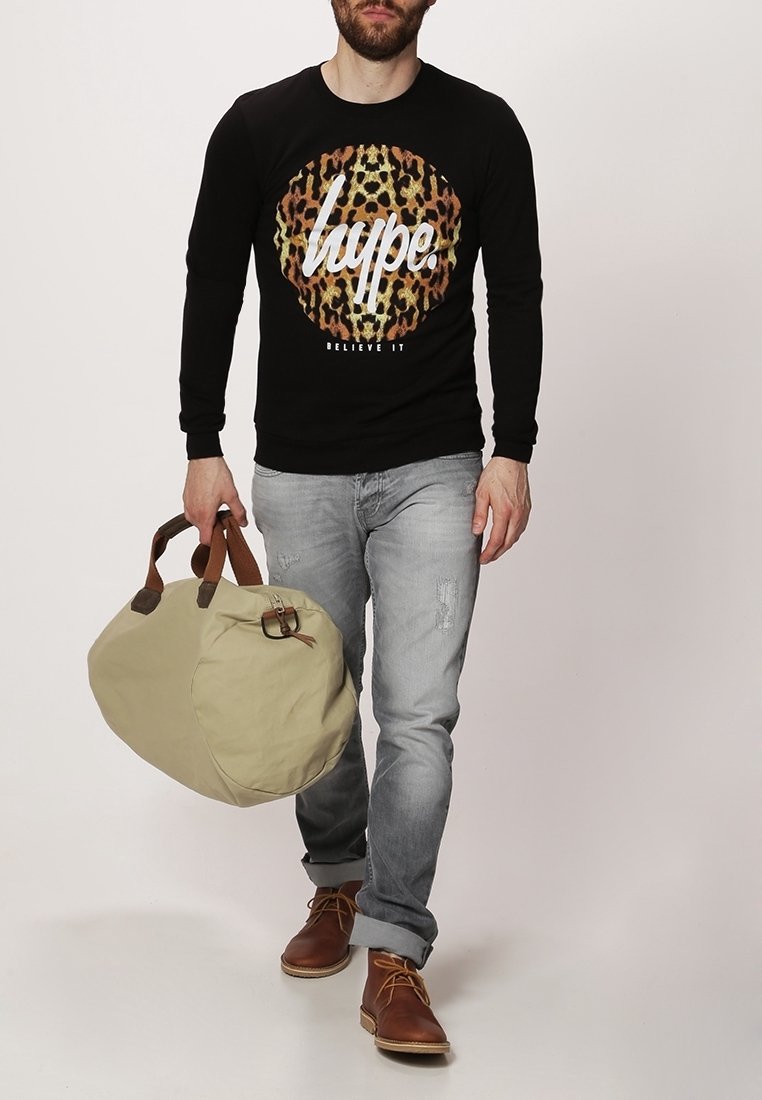 Homme portant une chemise noire à manches longues avec imprimé léopard et texte, jean gris clair, bottes marron, portant un sac polochon beige avec des poignées marron.
