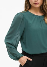 Blusa ligera en color verde azulado con mangas largas y abullonadas. Presenta un escote redondeado y detalles fruncidos en los hombros. Combinada con pantalones negros.