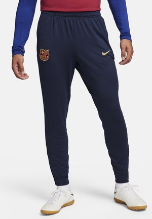 Pantalon de sport bleu marine avec écusson du FC Barcelone et logo Nike doré, porté avec des chaussures de sport blanches et un haut à manches longues bleu et rouge.