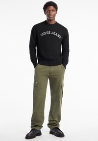 Černý pletený svetr s nápisem "GUESS JEANS", doplněný olivově zelenými cargo kalhotami s mnoha kapsami, zakončený černými botami.