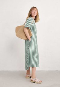 Abito a righe verde e bianco con maniche corte, dotato di bottoni e spacco laterale. La persona porta una borsa a tracolla intrecciata di colore beige e indossa sandali chiari.