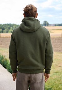 Olivgrüner Kapuzenpullover aus strukturiertem Stoff mit gerippten Bündchen. Der Pullover hat eine Kängurutasche und eine weiche Oberflächenbeschaffenheit.