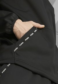 Chaqueta deportiva negra con un bolsillo tipo canguro; presenta un logotipo blanco de Puma a lo largo del borde del bolsillo; tela suave; puños elásticos.