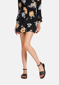 Tuta floreale nera con maniche corte e vita arricciata, abbinata a sandali neri con ritagli circolari e cinturino con fibbia.