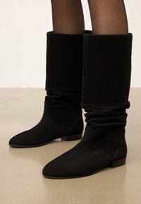 Juodos, laisvos kulkšnies botai, pagaminti iš minkštos medžiagos, su smailiu galiuku ir žemu blokuotu kulnu. Sklandus paviršius be matomų detalių.