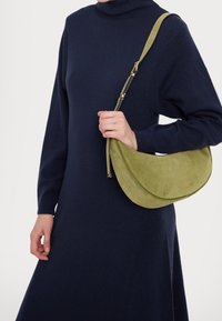 Grüne Wildleder-Handtasche mit geschwungener Form und goldfarbenen Beschlägen, über die Schulter getragen, kombiniert mit einem dunkelblauen Rollkragenkleid.