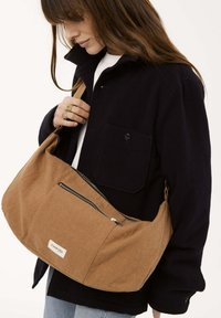 Sac à épaule en tissu brun avec une forme incurvée, un grand compartiment zippé et une petite étiquette. Porté sur un manteau sombre avec un design à col.