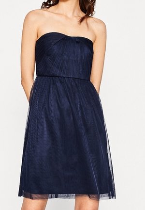 Femme portant une robe bleu marine sans bretelles, longueur genou, avec un corsage cintré et une jupe légèrement évasée.