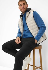 Gilet imbottito color beige chiaro sopra un felpa blu, pantaloni neri e sneakers bianche. Il gilet presenta un cappuccio e dettagli con zip.