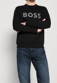 Sudadera negra con un destacado logo gris "BOSS" en la parte frontal. Presenta un cuello redondo y puños y dobladillo acanalados. Combinada con jeans azul oscuro.