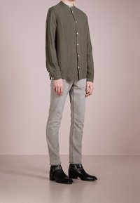 Olivově zelená košile s dlouhým rukávem a kulatým límcem, s knoflíkovým uzavřením a volným střihem, spárovaná s světle šedými úzkými džíny a černými kotníkovými botami.