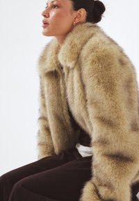 Falošná kožušina bunda v svetlo béžovej farbe so širokým golierom, dlhými rukávmi a jemnou, textúrovanou povrchovou úpravou. Nosená cez tmavý outfit.