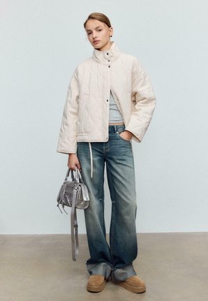 Kvinde iført lys off-white quiltet jakke, lysegrå cropped top, vidbenede, falmede jeans, tanfarvede ruskindssko og holding en sølvfarvet håndtaske mod en ensfarvet baggrund.