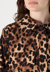Camicia con stampa leopardata marrone, colletto bottone, bottoni neri e taschino sul petto, realizzata in tessuto morbido con una superficie liscia.