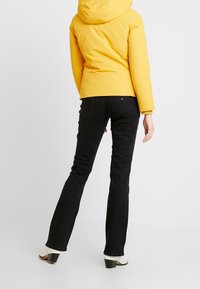 Geel geïsoleerde jas met een capuchon, gecombineerd met zwarte klokvormige jeans. De jeans hebben een klassiek vijf-pocketontwerp en een subtiele ruglogo.