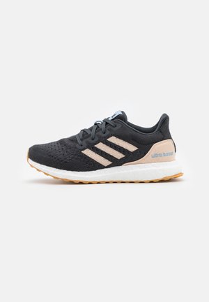 Chaussure de running Adidas Ultra Boost noire et beige avec empeigne en tricot, semelle blanche amortissante, semelle extérieure en gomme et trois bandes beiges sur le côté.