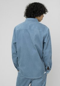 Chemise en velours côtelé bleu clair à manches longues, col classique et texture côtelée verticale. Dotée d'ouvertures latérales et de poignets boutonnés.