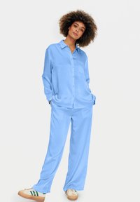 Donna con i capelli ricci che indossa una camicia blu chiaro sbottonata e pantaloni abbinati, in piedi con le mani nelle tasche e scarpe da ginnastica beige.