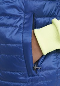 Niebieska pikowana kamizelka o gładkiej, błyszczącej fakturze. Posiada pionową, zapinaną na zamek boczną kieszeń oraz ściągacze w jasnym żółtym kolorze.