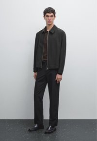 Giovane uomo in piedi con giacca nera con cerniera, camicia marrone scuro, pantaloni neri e mocassini neri contro una parete grigia chiara uniforme.