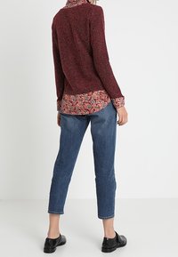 Maglione rosso testurizzato indossato sopra una camicia a fiori, abbinato a jeans blu a sigaretta e scarpe nere. Caratteristiche includono polsini con bottoni.