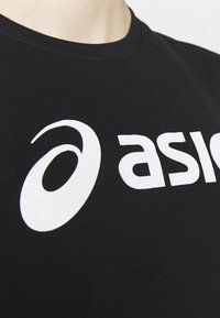 Nahaufnahme eines schwarzen T-Shirts mit einem weißen Asics-Logo und teilweise auf der Brust aufgedrucktem Text.