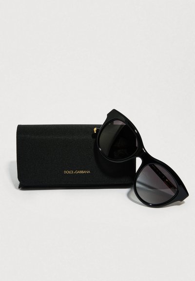 Juodos katės akių formos akiniai nuo saulės su blizgiu paviršiumi ir tamsiais lęšiais, padėti šalia tekstūruotos juodos dėžutės su auksiniu prekės ženklu.