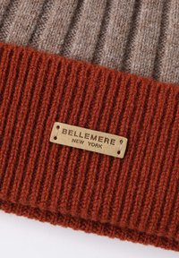Berretto lavorato a maglia con parte superiore marrone testurizzata e fascia a coste spessa di colore ruggine. Presenta un'etichetta rettangolare beige impressa con la scritta "Bellemere New York."