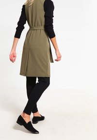 Vestido sin mangas color verde oliva con cintura ceñida, combinado con leggings negros y zapatos slip-on negros. Tela suave y ajuste entallado.