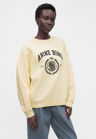 Γυναίκα φορώντας ένα oversized ανοιχτό κίτρινο φούτερ με το λογότυπο «ANINE BING» και ανοιχτό μπλε άνετο τζιν, στέκεται μπροστά σε απλό λευκό φόντο.