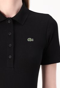 Juodas raštuotas polo marškinėlis su smailiu apykakle, mygtukų priekiu ir žaliu siuvinėtu krokodilų logotipu kairiajame krūtinės kampe.