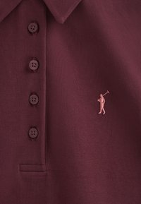 Bordowa koszulka polo z fakturowanej tkaniny, z sześcioma dopasowanymi guzikami i małym różowym golfistą haftowanym po lewej stronie klatki piersiowej.