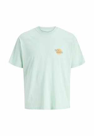 Uśmiechnięty chłopiec stoi przodem, ubrany w jasnografitowy t-shirt, ciemne spodenki do kolan, białe skarpetki i białe trampki na jednolitym tle.