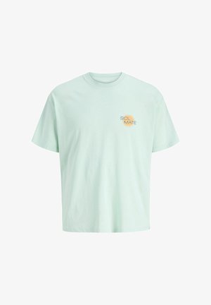 Smilende dreng står vendt fremad iført en lysegrøn t-shirt, mørke knælange shorts, hvide sokker og hvide sneakers på en simpel baggrund.