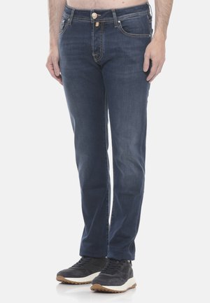 Uomo che indossa jeans slim fit blu scuro e sneakers nere con suola bianca, in piedi davanti a uno sfondo neutro.