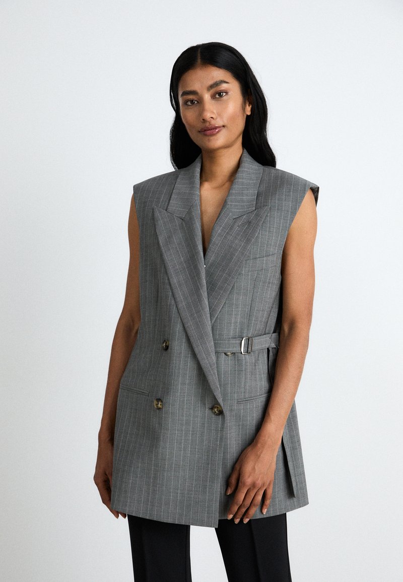 Nainen, jolla on pitkät mustat hiukset, pukeutuu ilmanhihaisempaan harmaaseen raidoitettuun bleiseriin, jossa on sivuhihna, poseeraa yksinkertaista vaaleaa taustaa vasten.