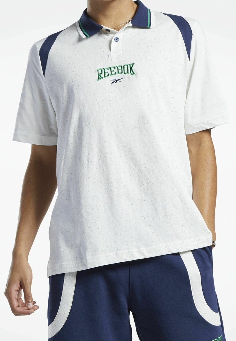 Homme portant un polo blanc Reebok avec des accents bleu marine et vert, assorti à un short bleu marine avec des bandes blanches incurvées sur les poches.