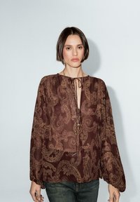 Blusa marrone paisley con colletto a nodi, lunghe maniche ampie e una consistenza morbida e traslucida. Abbinata a jeans verde scuro.