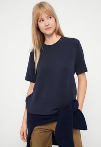 Νεαρή γυναίκα με ξανθά μαλλιά, φορώντας ένα ναυτικό μπλε μπλουζάκι κοντό μανίκι και μια ταιριαστή ζακέτα δεμένη γύρω από τη μέση της, μαζί με καμηλό παντελόνια.