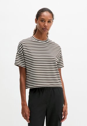 Femme aux cheveux bouclés foncés attachés, portant un t-shirt à manches courtes rayé horizontal noir et blanc et un pantalon noir sur un fond uni.