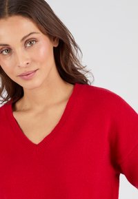 Maglione rosso a maglia con scollatura a V, caratterizzato da una texture morbida e una vestibilità rilassata, con dettagli a coste visibili sulla scollatura e sulle maniche.