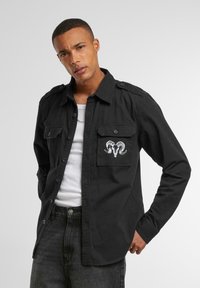 Black button-up shirt with a pocket, featuring a white ram skull embroidery. Made of cotton, it has a relaxed fit and a pointed collar.