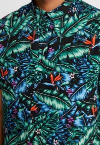 Kurzarmhemd mit schwarzem Hintergrund und lebhaften tropischen Blattmustern in Grün und Blau, akzentuiert durch orangefarbene und lila Blumen.