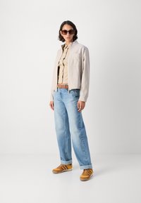 Giacca cropped beige chiaro, camicia button-up color sabbia, jeans a gamba larga blu chiaro e sneakers marroni con dettagli gialli. Occhiali da sole marroni e orecchini.