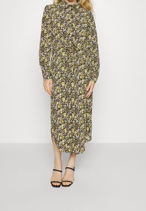 Femme portant une robe midi à manches longues avec un motif floral de petites fleurs jaunes et violettes, associée à des sandales noires à talons et à brides.