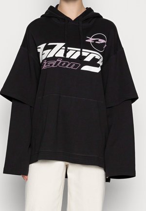 Osoba ubrana w czarną, oversize'ową bluzę z kapturem z długimi rękawami nałożonymi pod spodem, z białym i fioletowym nadrukiem tekstu i logo na piersi, w połączeniu z jasnymi spodniami.
